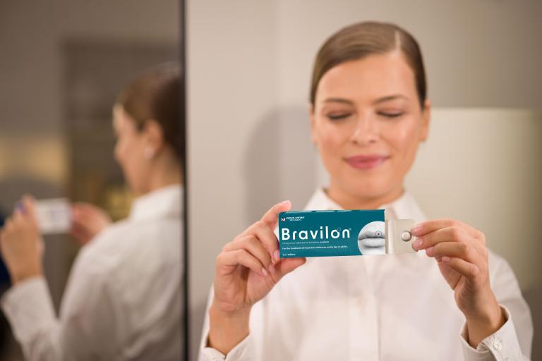 Žena drží otevřené balení přípravku Bravilon® s viditelným blistrem tablet, mírně se usmívá a má spokojeně přivřené oči.